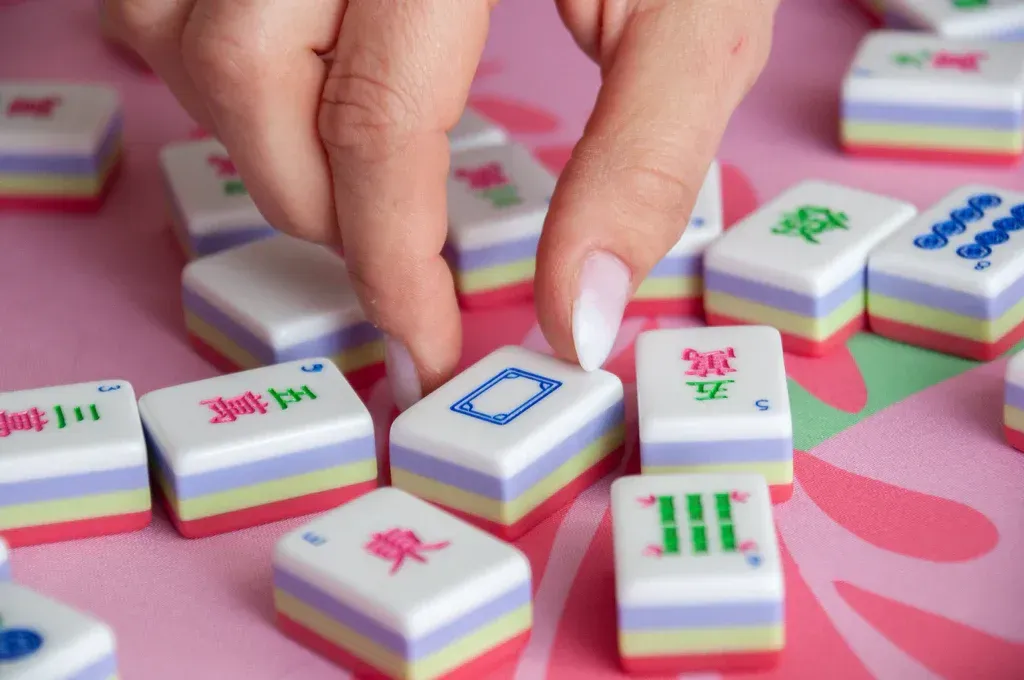Hand picking up a Mahjong tile from a table. Tiles are white with red, green, and blue symbols. Pink table.