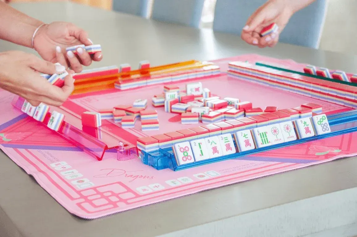 People playing Mahjong on a pink table. Hands are holding tiles. Close-up shows the game setup with tiles.