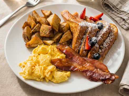 A breakfast plate with scrambled eggs, crispy bacon, home fries, and French toast topped with berries and powdered sugar.