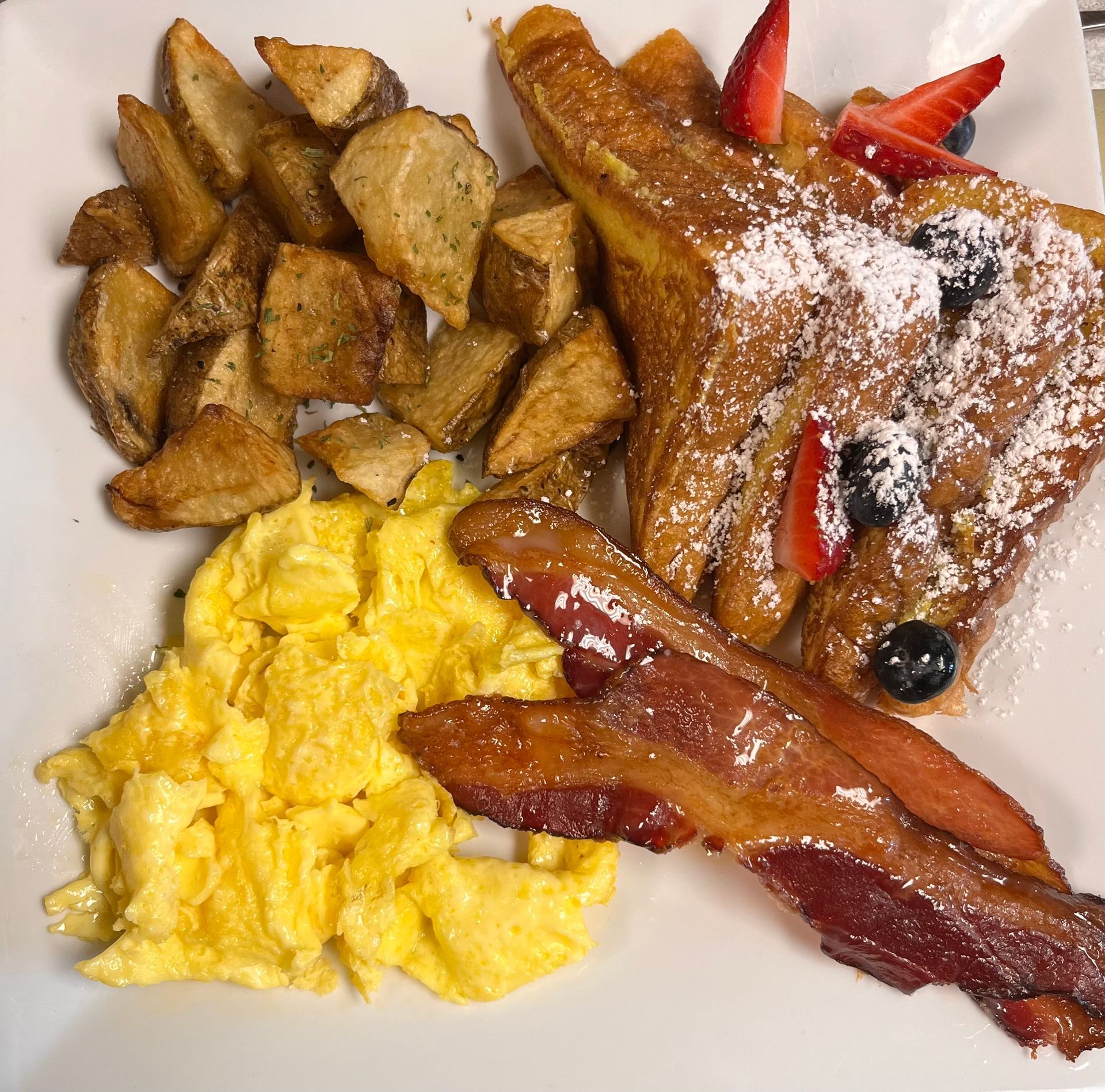 A breakfast plate with scrambled eggs, crispy bacon, home fries, and French toast topped with berries and powdered sugar.