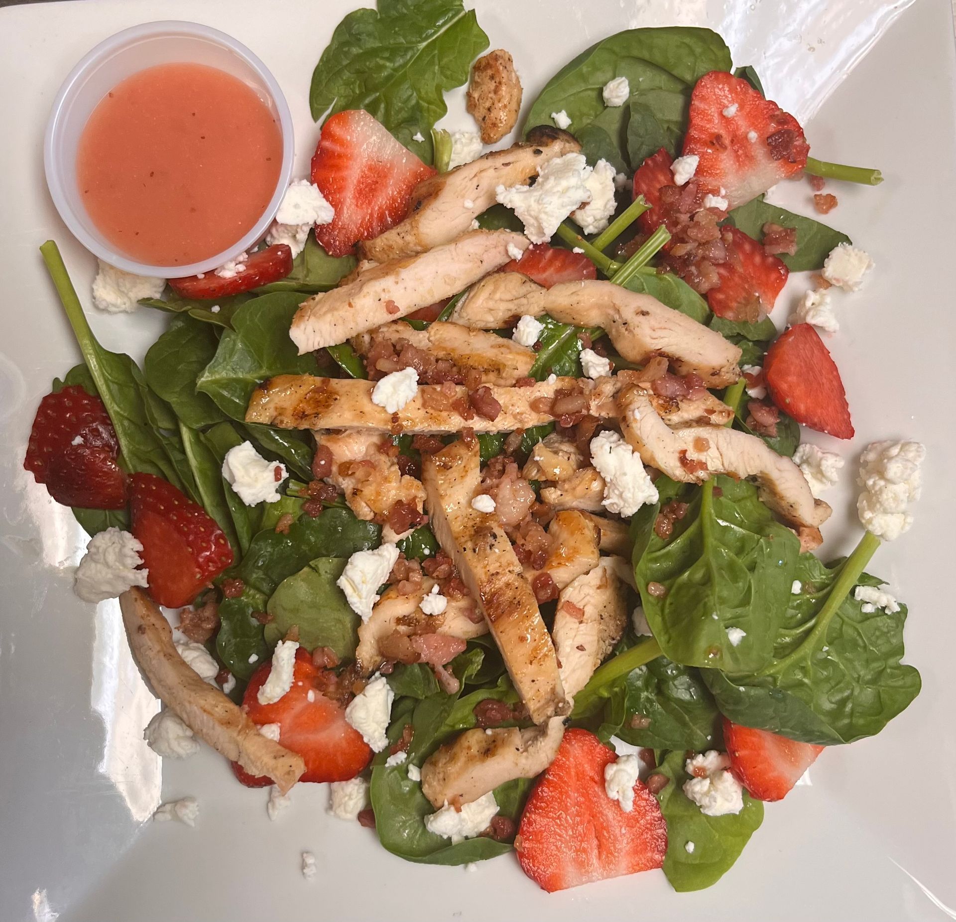 A spinach salad topped with grilled chicken, sliced strawberries, crumbled white cheese, and a small cup of dressing.