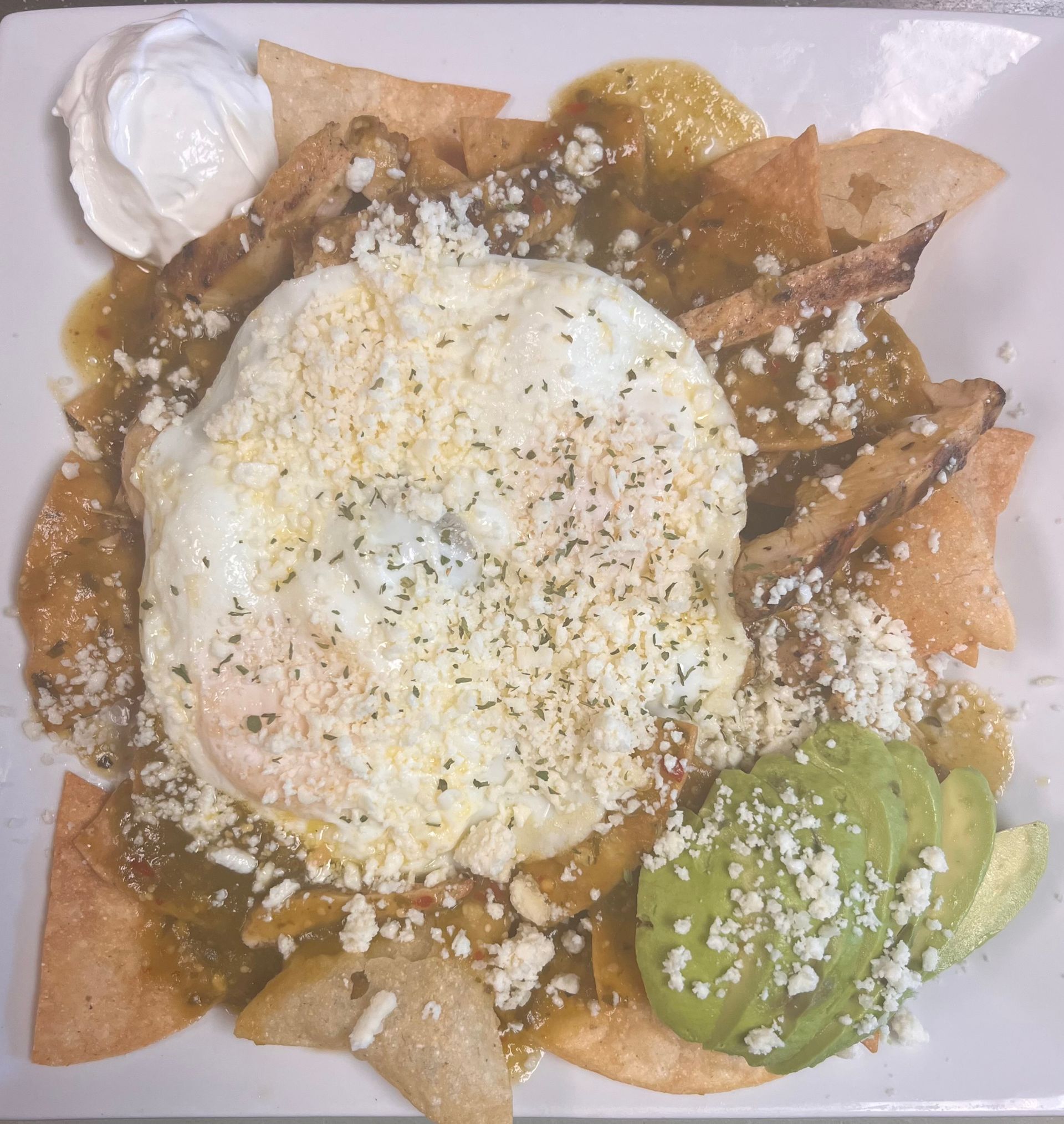 A plate of chilaquiles with green salsa, a fried egg, crumbled cheese, sour cream, and avocado slices on corn chips.