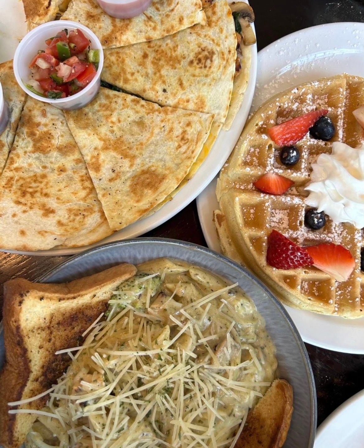 Three plates of food: quesadillas with salsa, a waffle with berries and whipped cream, and pasta with toast.