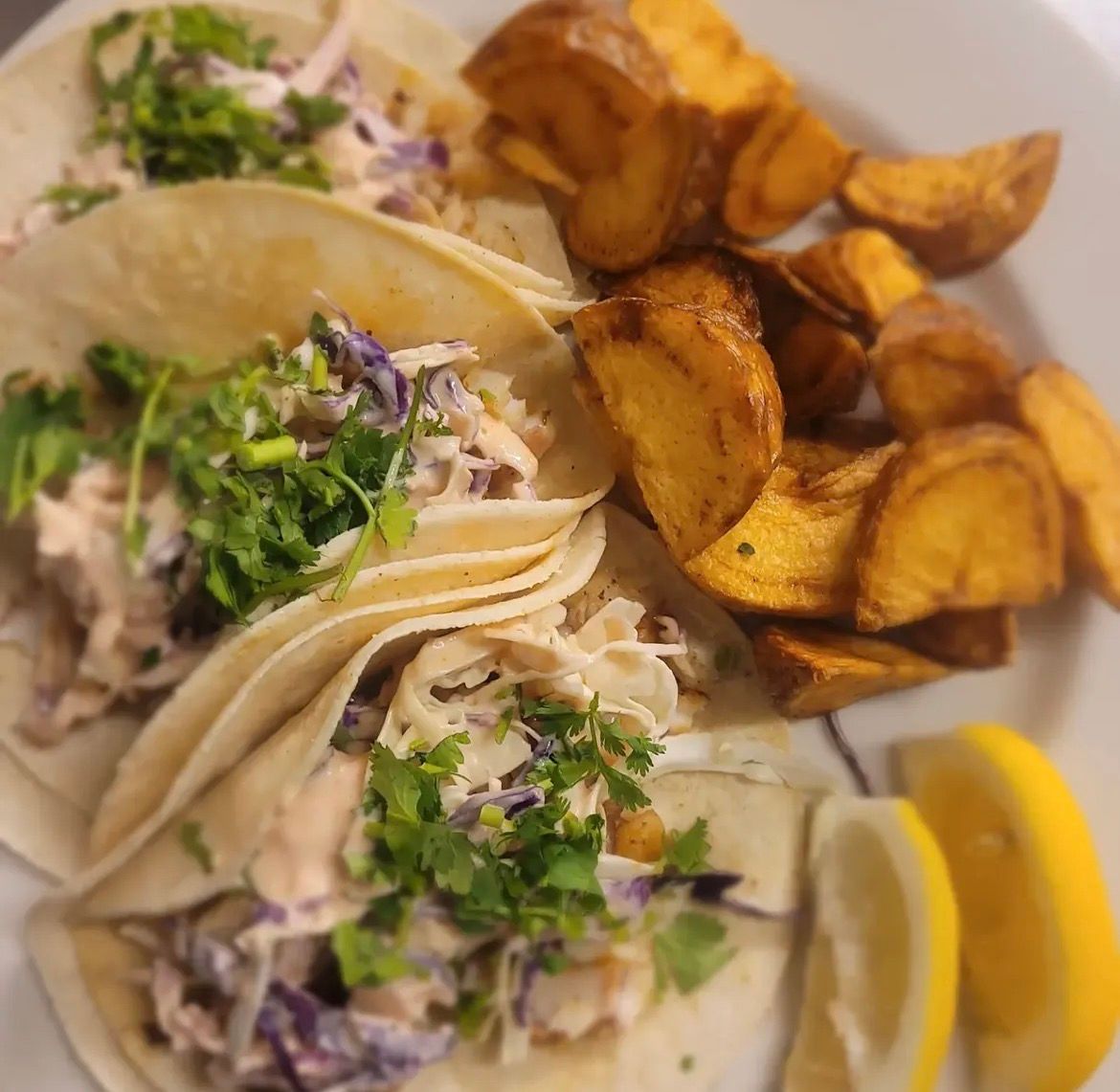 Three fish tacos topped with slaw and cilantro, served with seasoned potato wedges and lemon slices on a white plate.