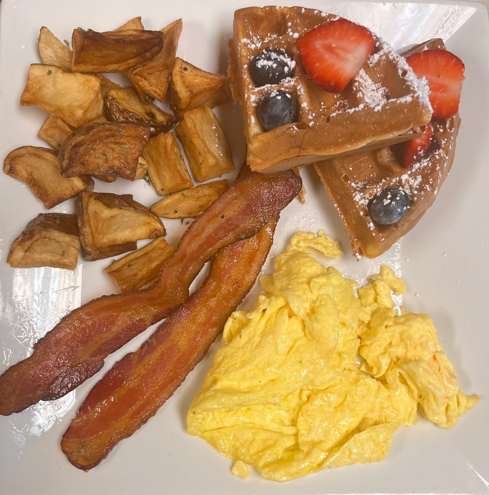 A breakfast plate with scrambled eggs, two strips of bacon, seasoned potato wedges, and waffles topped with berries.