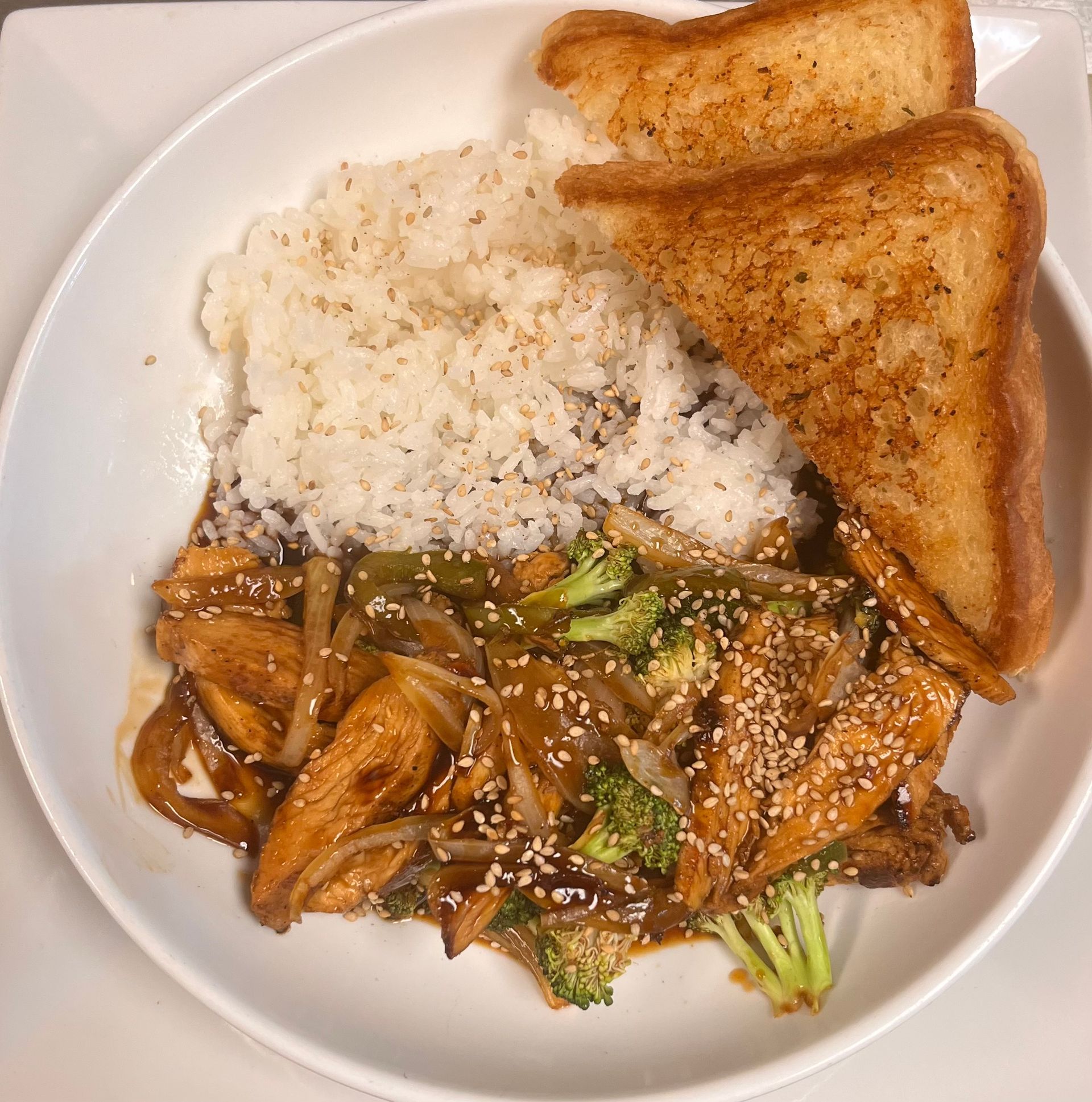 A white bowl containing chicken and broccoli stir-fry with a side of white rice and two pieces of toasted bread.