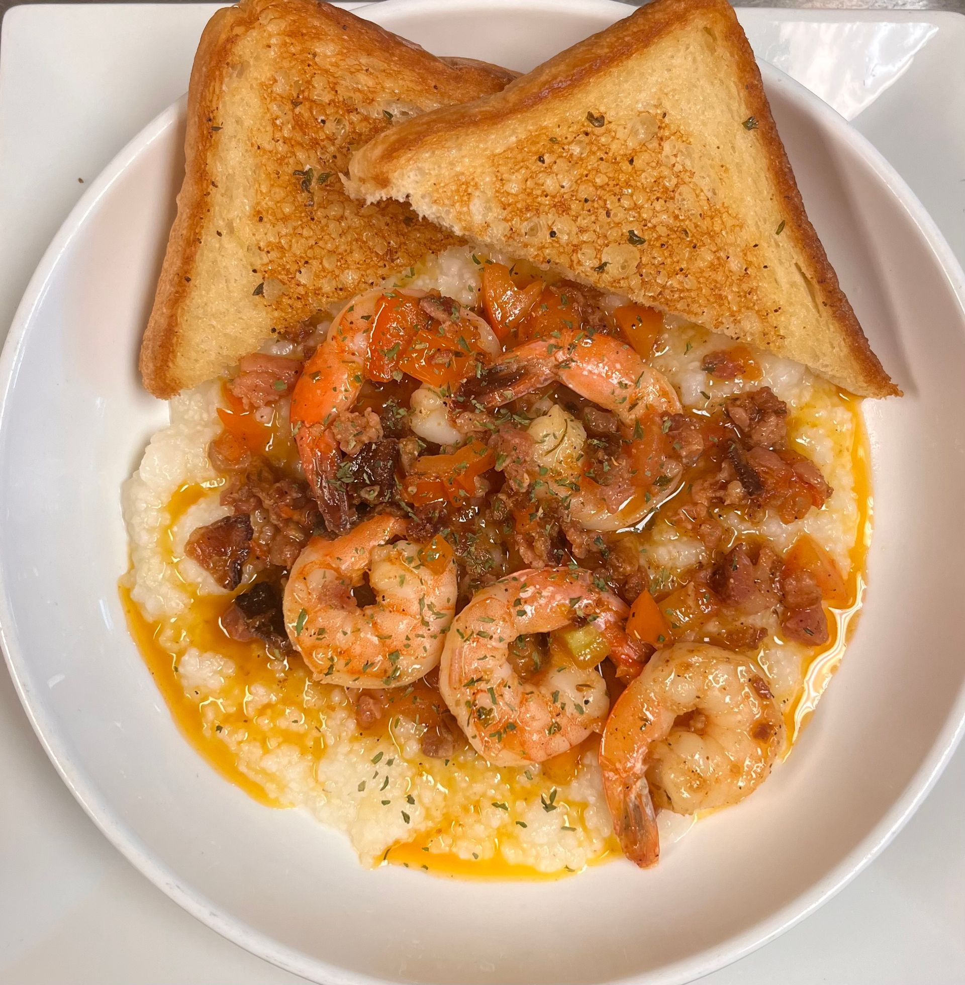 A bowl of shrimp and grits topped with bacon and a flavorful sauce, served with two pieces of toasted bread.