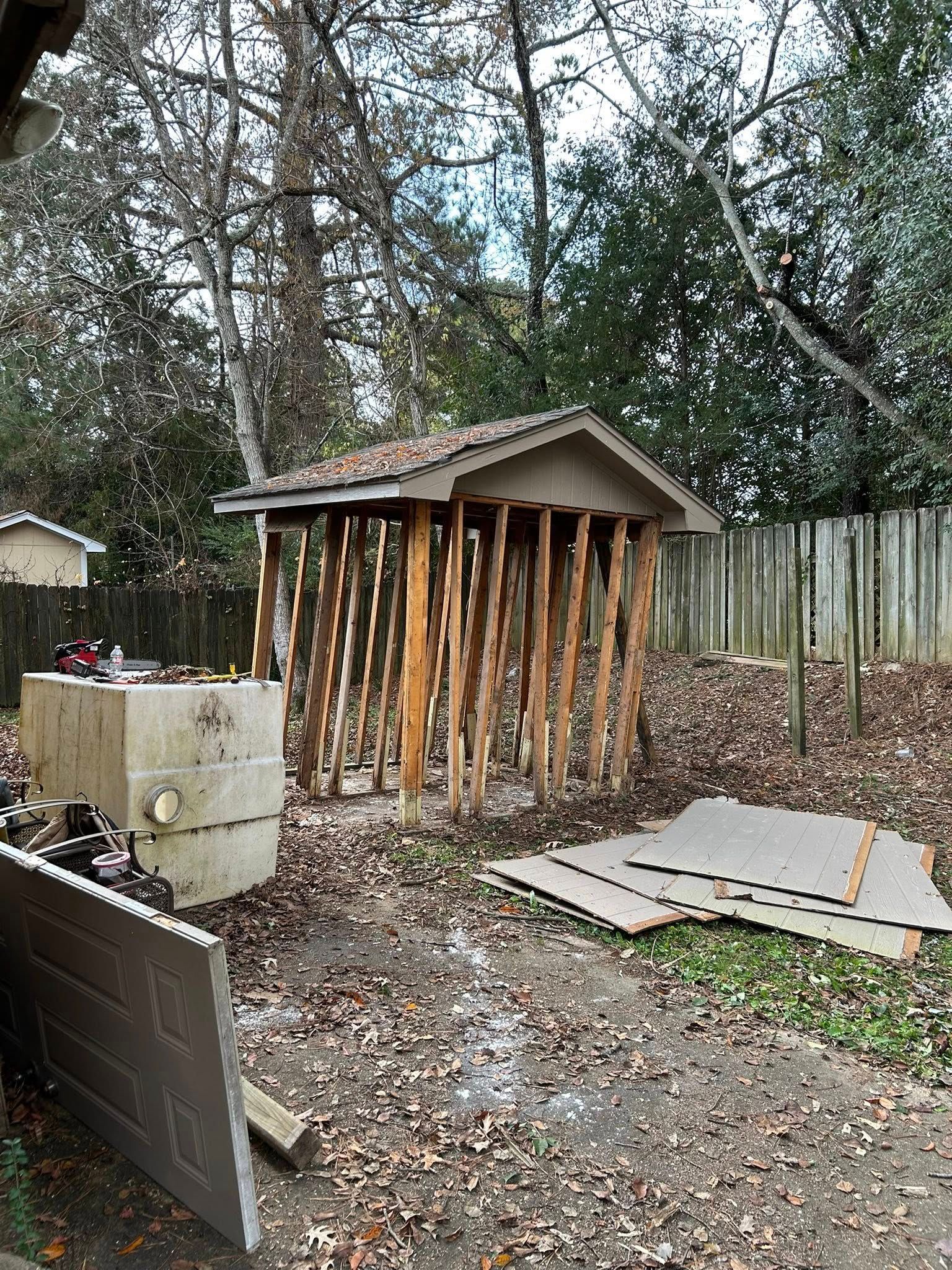 Small shed with exposed wooden supports in a muddy backyard, with fallen leaves and a fence behind it.