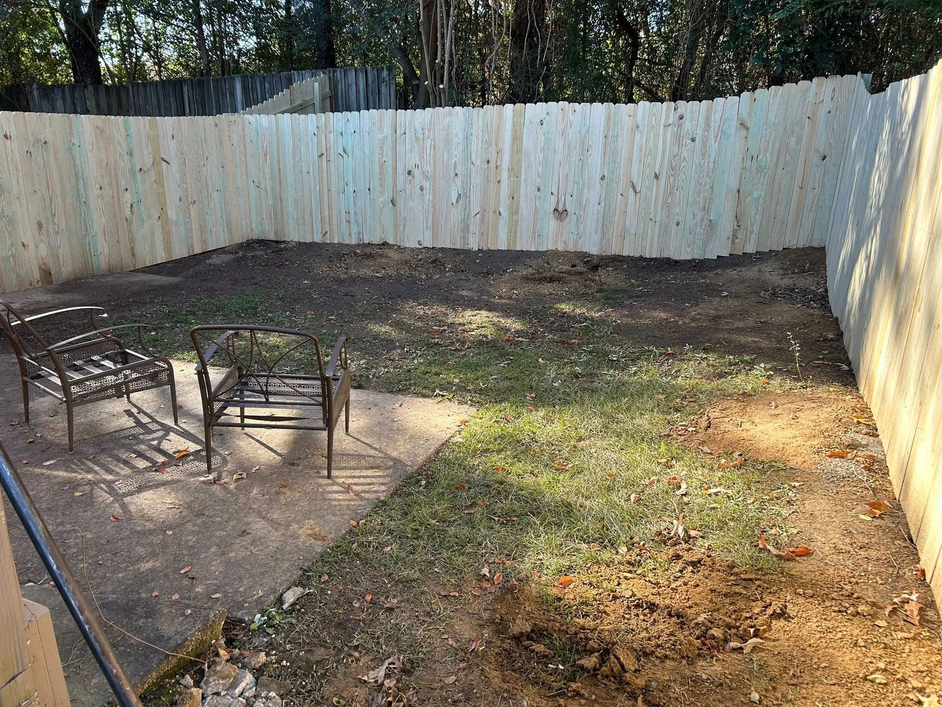 Empty fenced backyard with patchy grass, concrete patio, and two metal chairs near the left side.