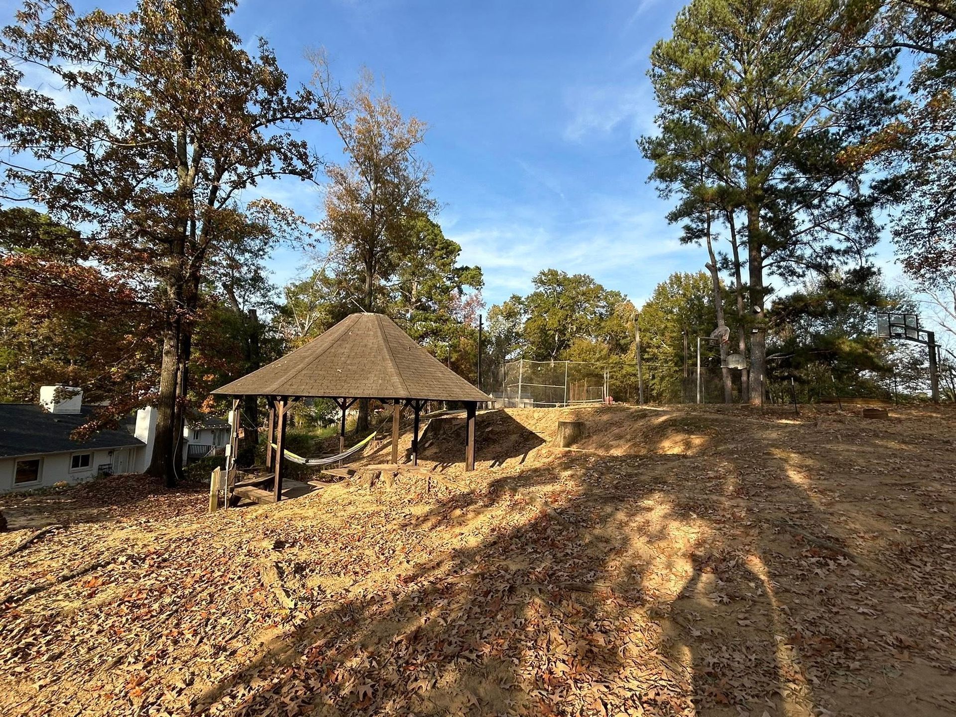 Wooded outdoor area with a thatched pavilion and a dirt hillside under a blue sky