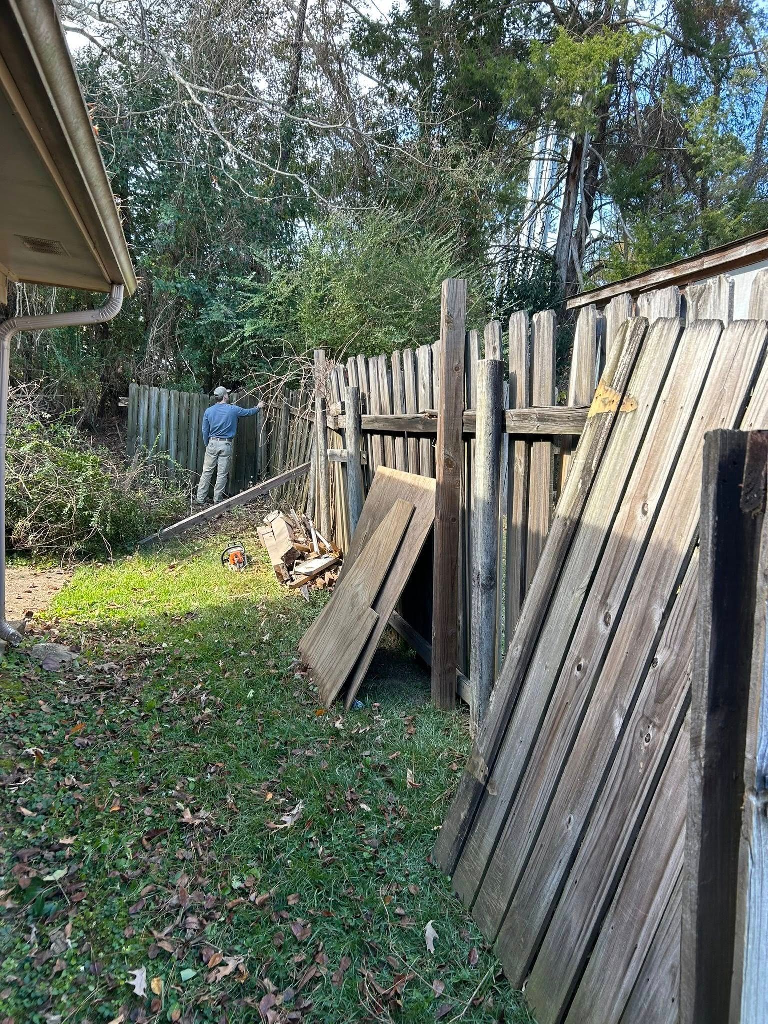 Person working near a leaning wooden fence beside a house in a leafy yard