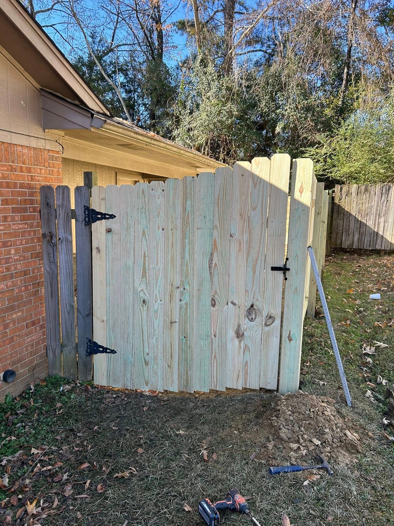 Weathered wooden gate with black hinges beside a brick house, in a grassy yard with trees behind it.