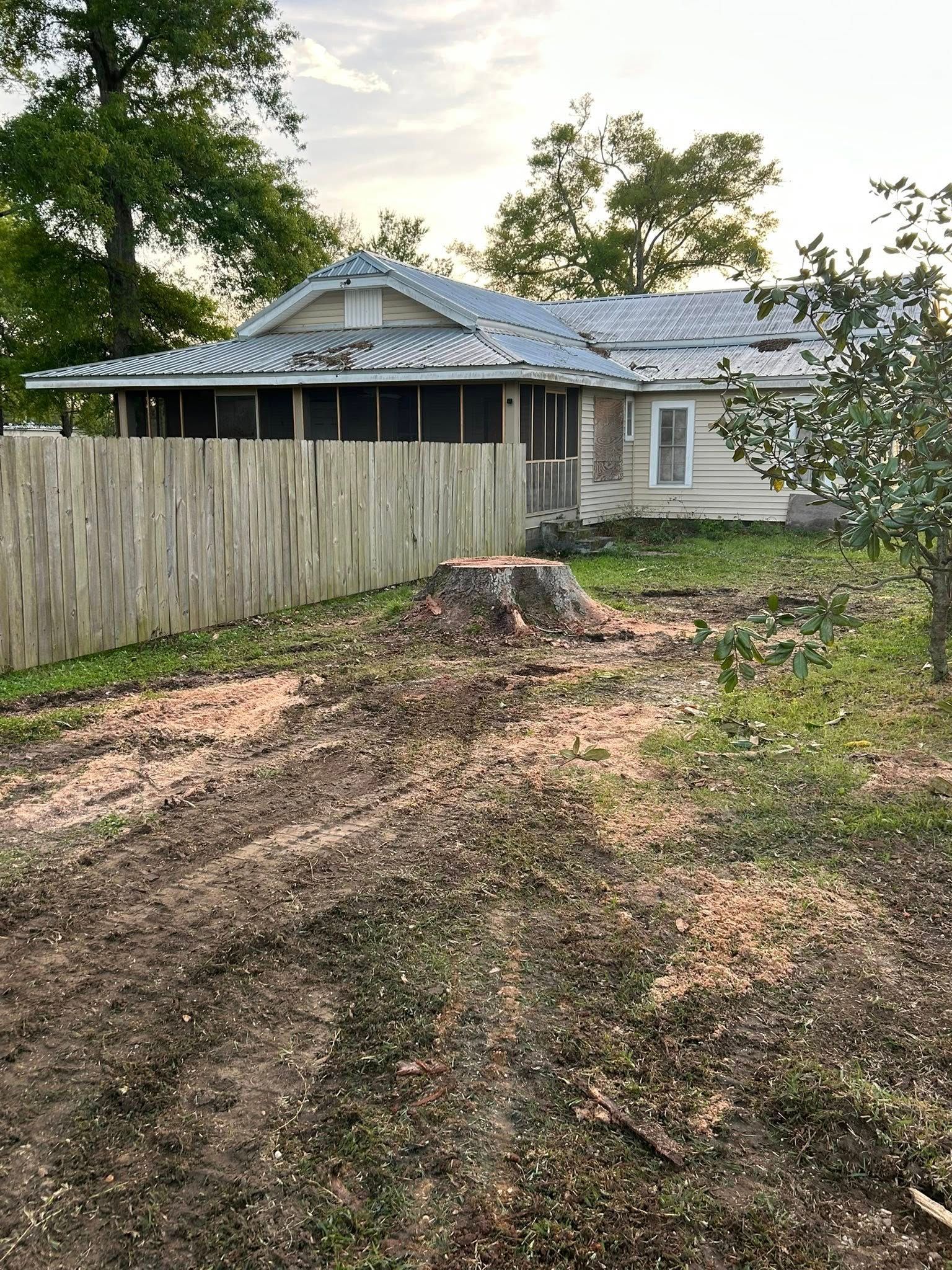 Rural house with damaged roof, leaning fence, and large tree stump in a muddy yard