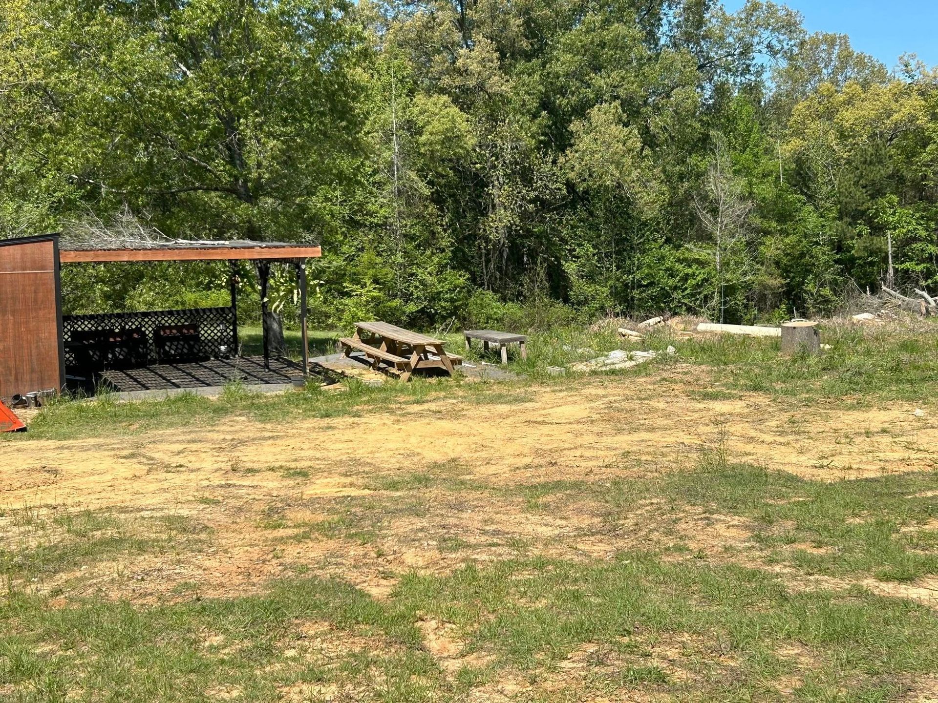 Grassy yard with a shaded shelter, picnic table, and trees in the background.