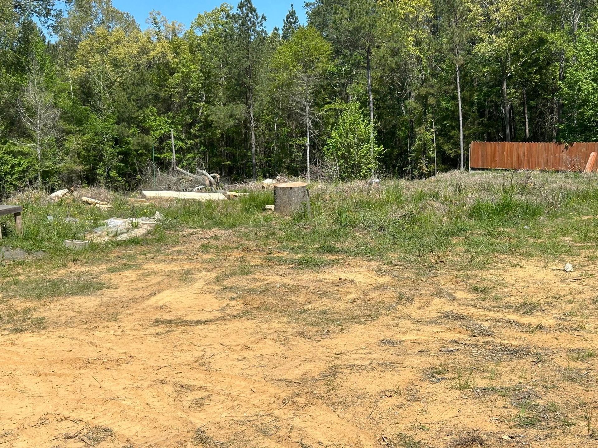 Dirt clearing beside a wooded area, with a rusty fence and blue sky in the background