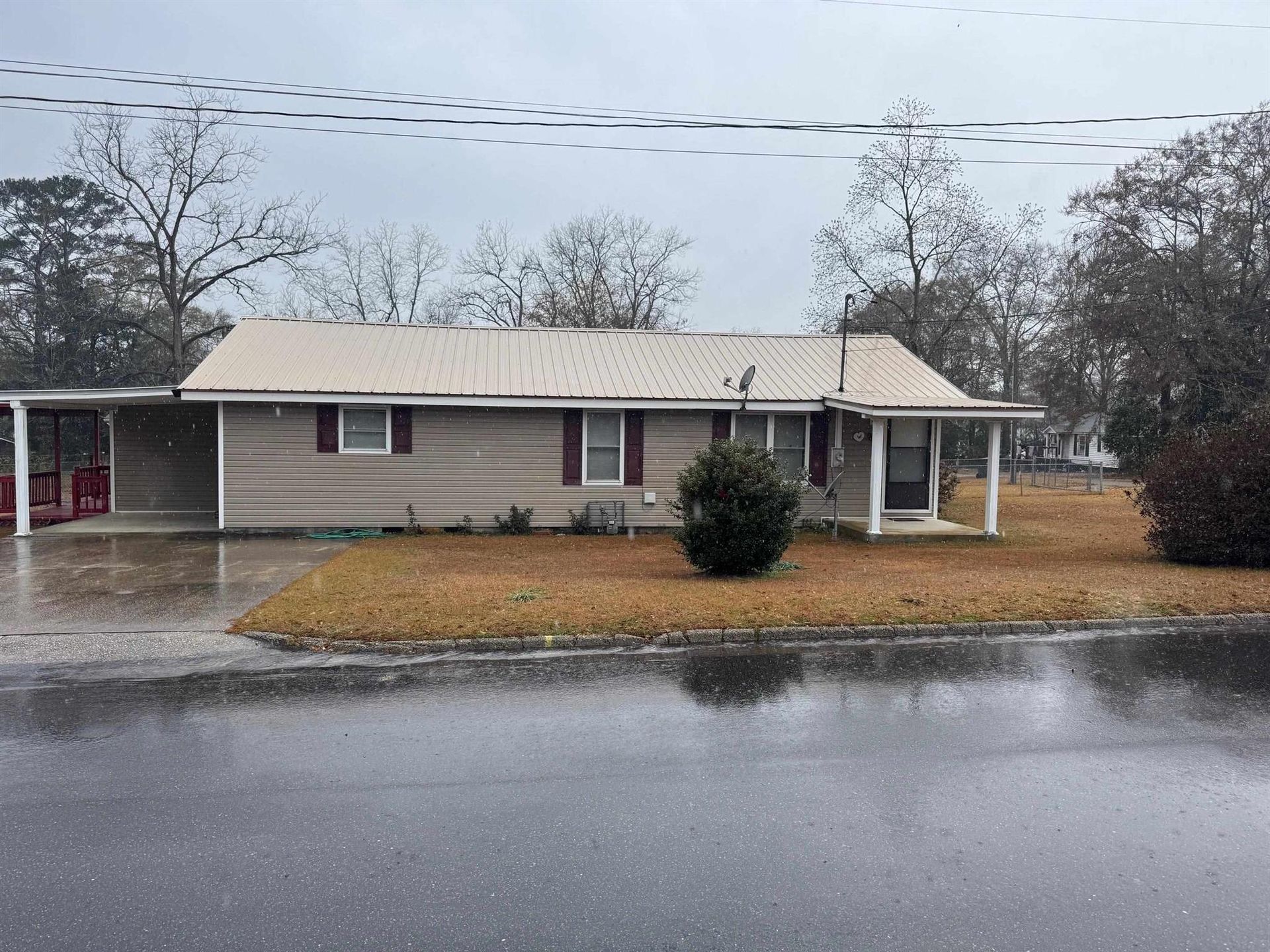 Tan house with carport and covered porch on a gray day. Metal roof, bare trees, wet street.