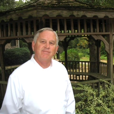 Man in white shirt in front of a wooden gazebo in a garden setting.