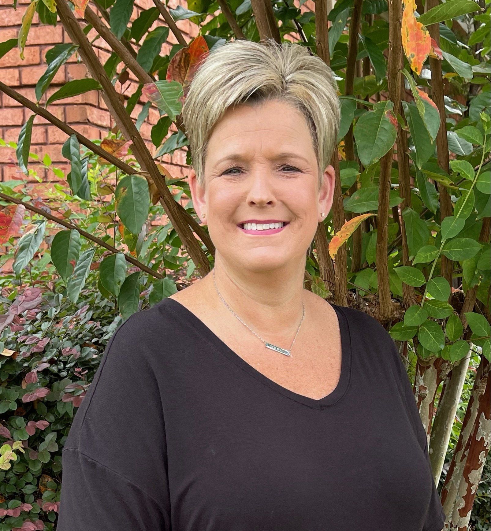 Woman with short blonde hair and a black top smiles in front of green and brick background.