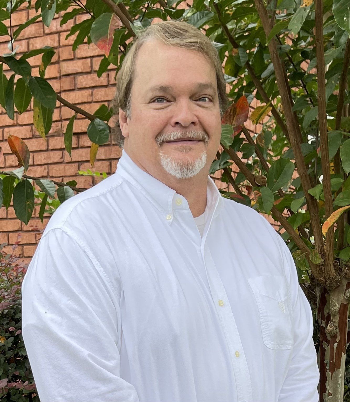 Man with light hair and goatee in a white shirt stands outdoors in front of a brick building.