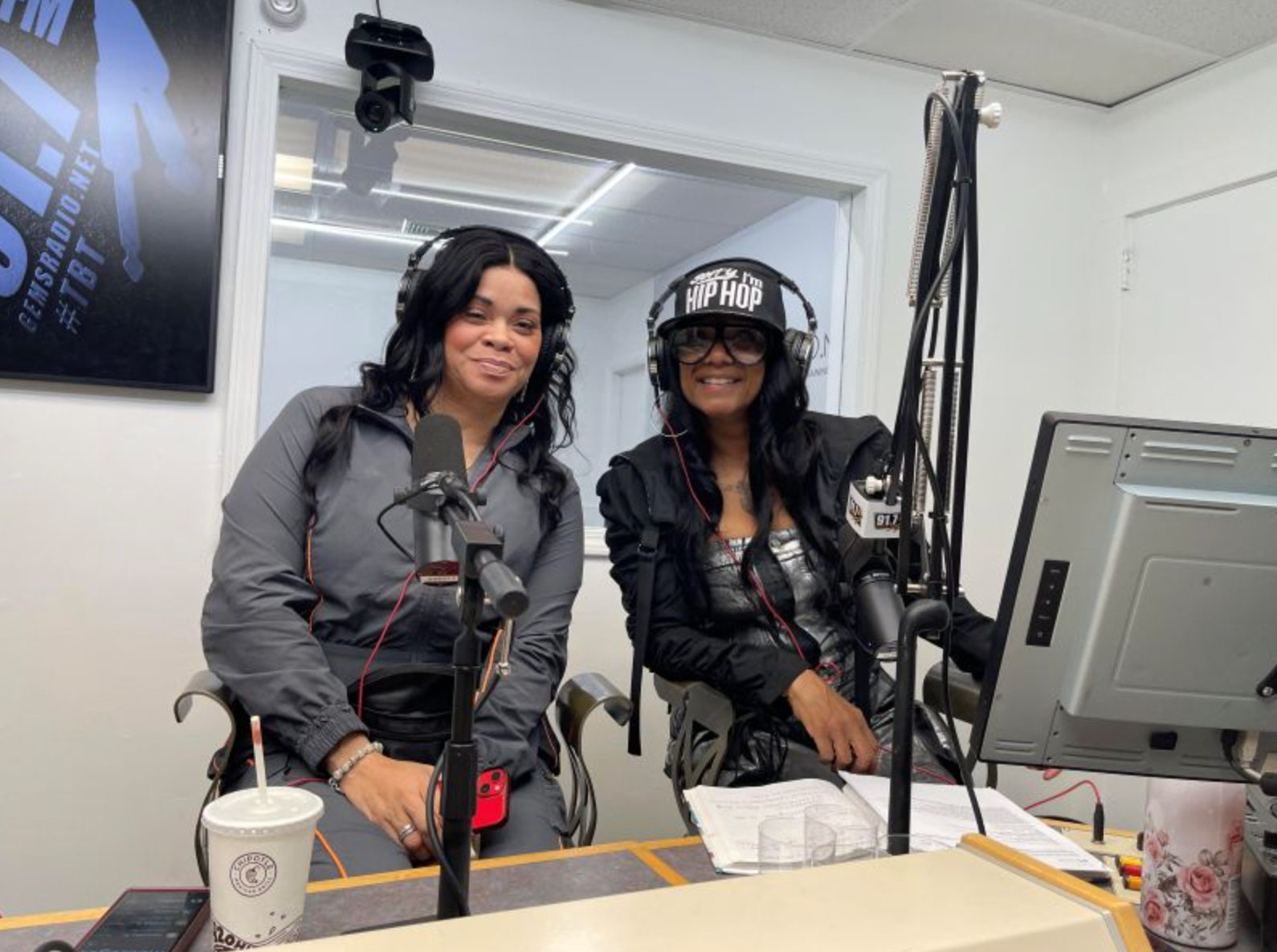 Two women in a radio studio at mics. One smiles, the other wears a helmet-like head covering.