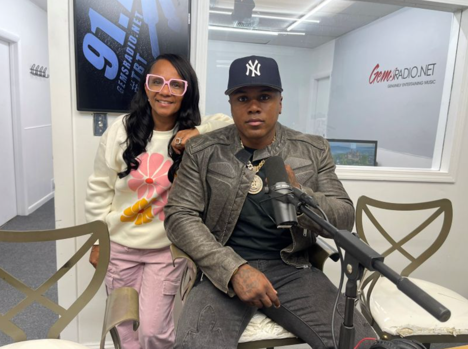 Woman and man in a radio studio. Woman wears pink glasses, man in Yankees cap. Both smiling near a microphone.