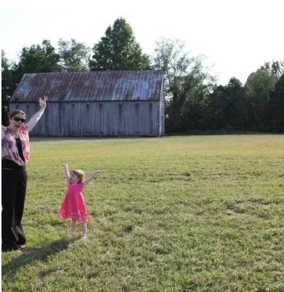 A woman and a little girl are standing in a field with a barn in the background