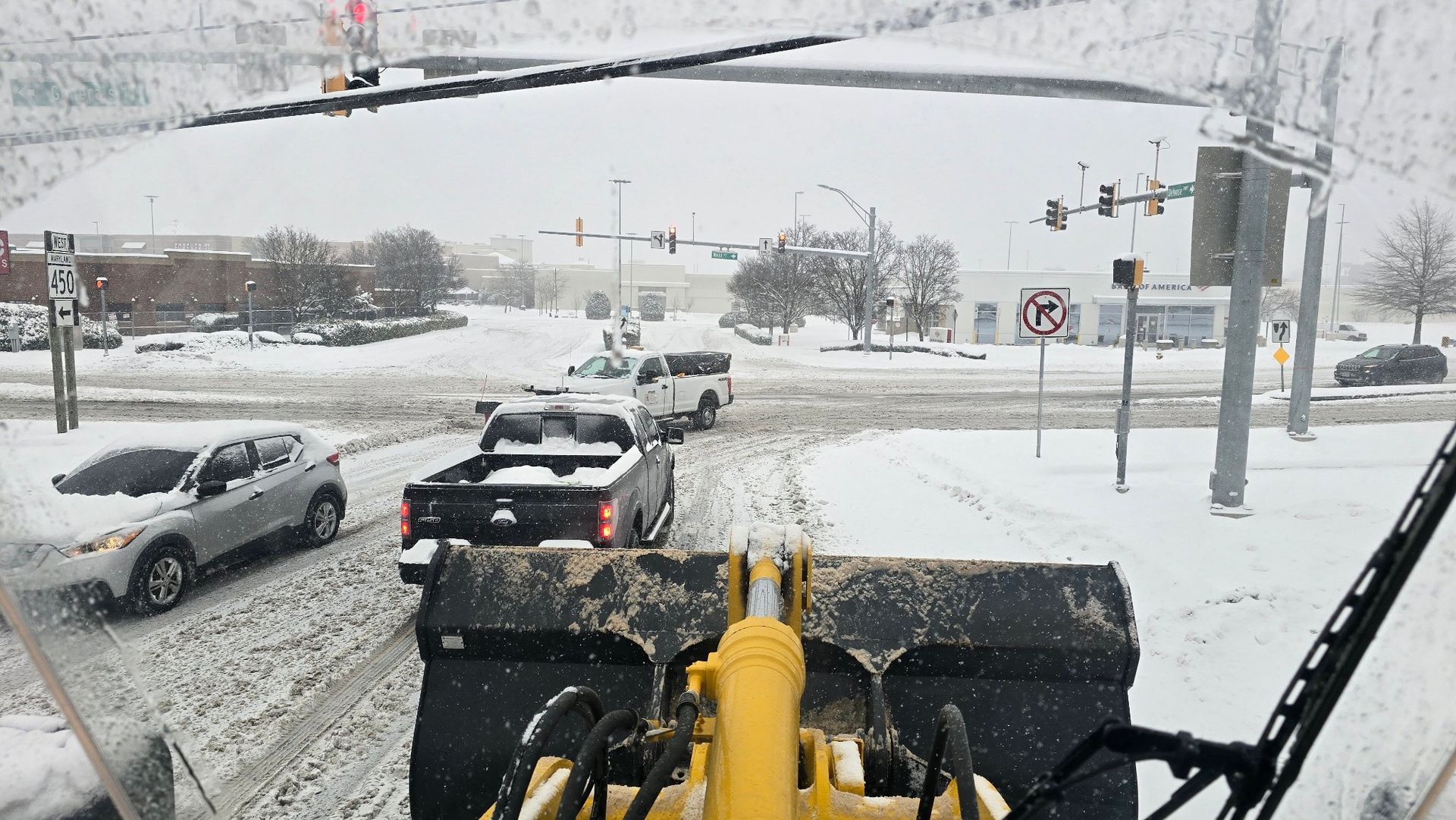 A snowplow clearing a snow-covered intersection with cars stopped nearby.
