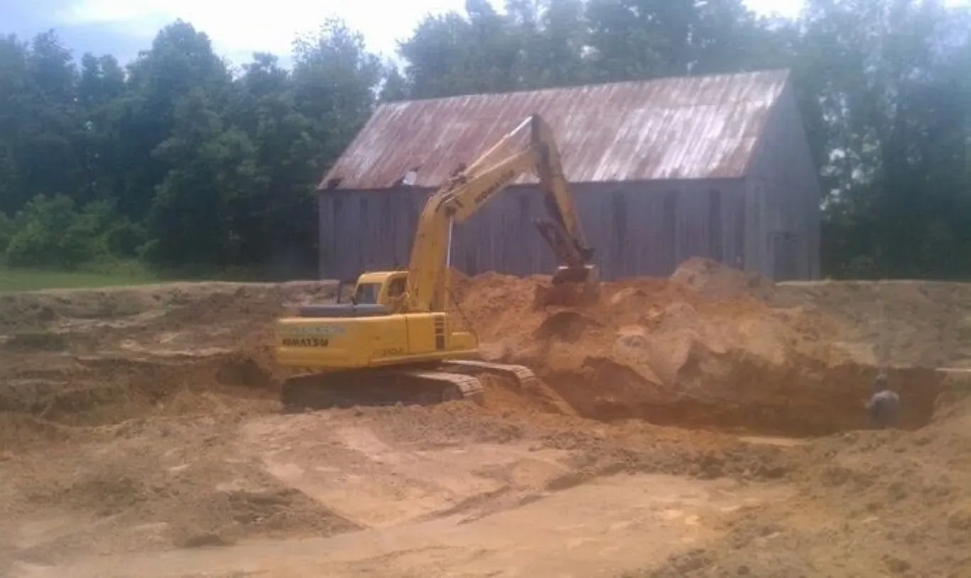 A yellow excavator is digging a hole in the dirt in front of a barn.