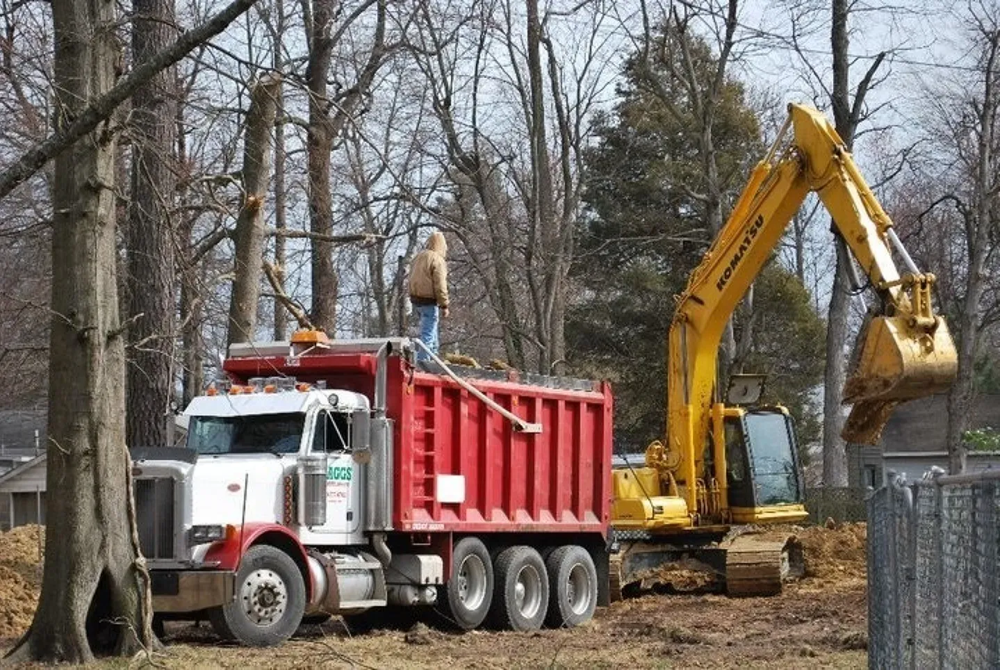 A man stands on top of a dump truck next to a yellow excavator