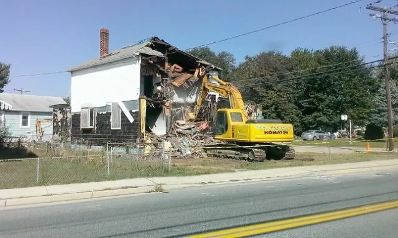 A yellow excavator is demolishing a house on the side of the road. 