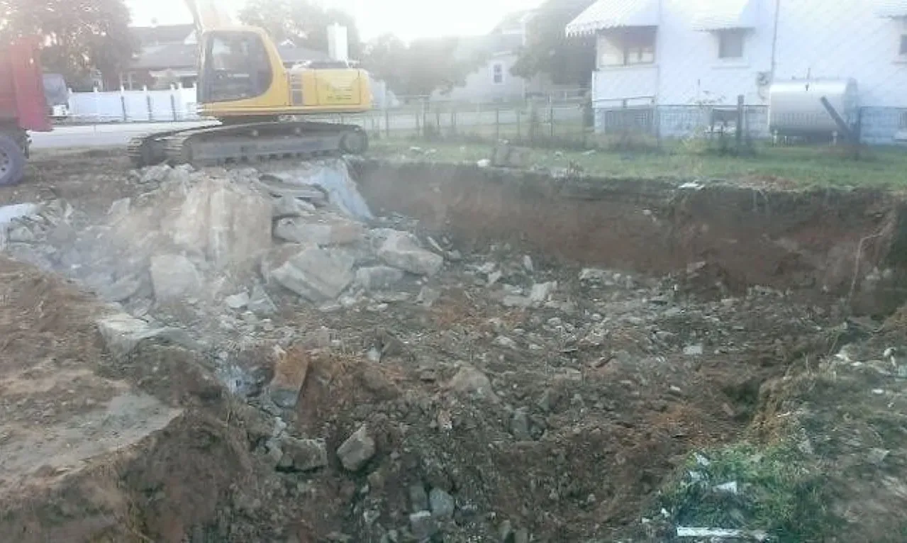 A yellow excavator is digging a hole in the dirt in front of a house.