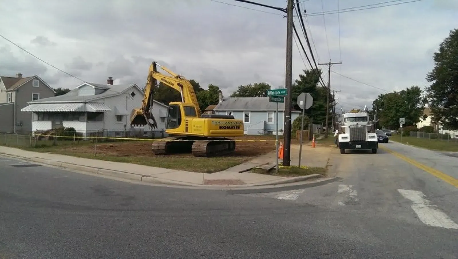 A yellow excavator is sitting on the corner of a street next to a truck.