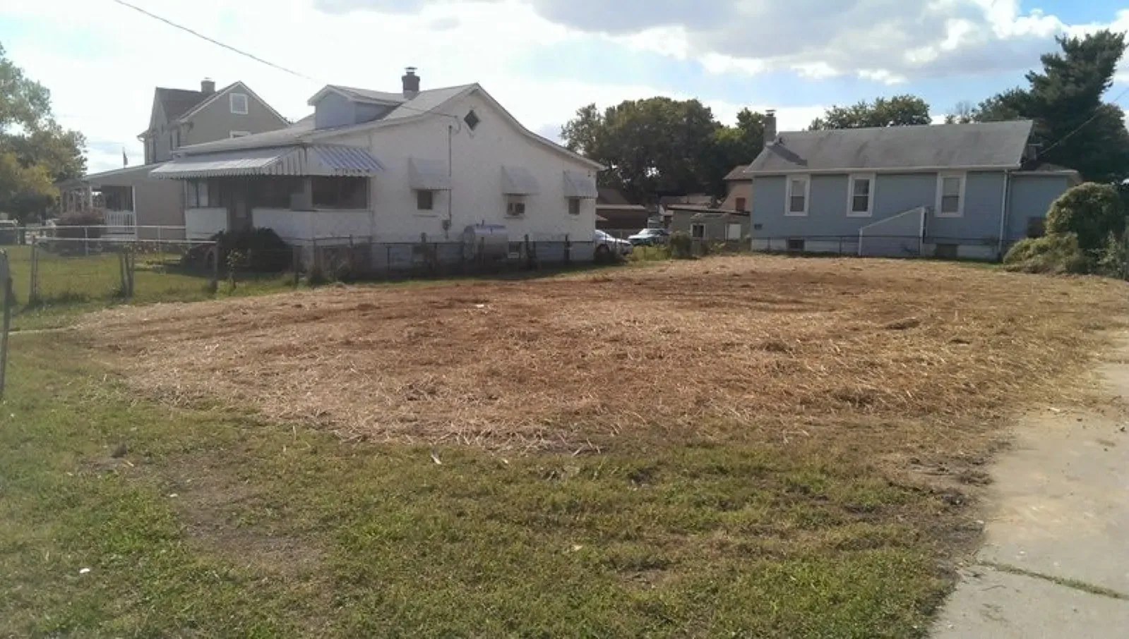 A white house is sitting in the middle of a grassy field next to a cleared lot ready to be built on. 
