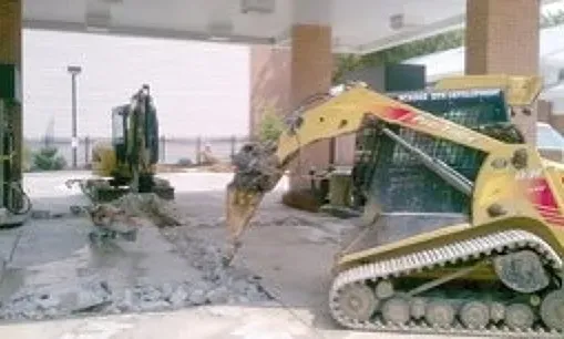 A yellow excavator is digging a hole in a parking lot.