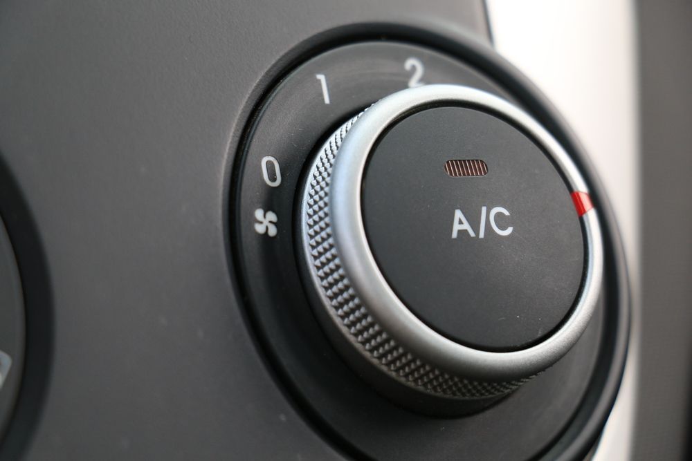 Close-up of a car's air conditioning dial with numbers 0, 1, 2 and A/C button, knob is silver, background is gray.