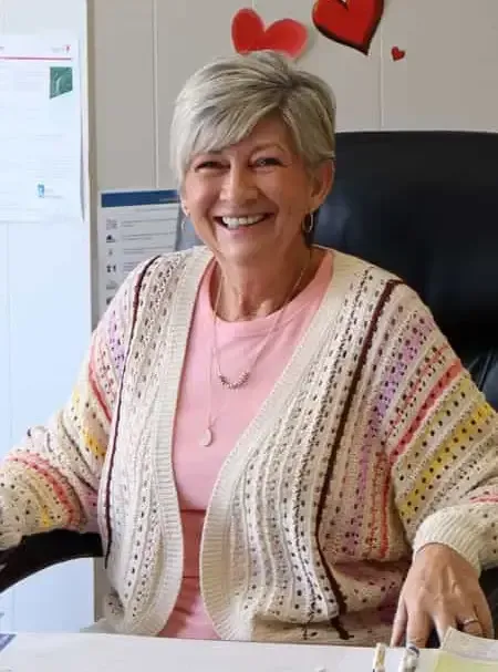 Smiling woman in a pink shirt and patterned cardigan seated at a desk with heart decorations behind her