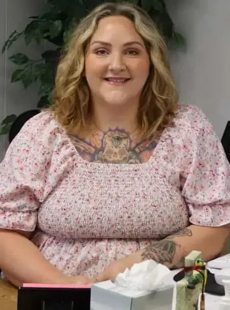 Smiling woman in a pink floral top seated indoors, with tattoos visible and a tissue box on the table.