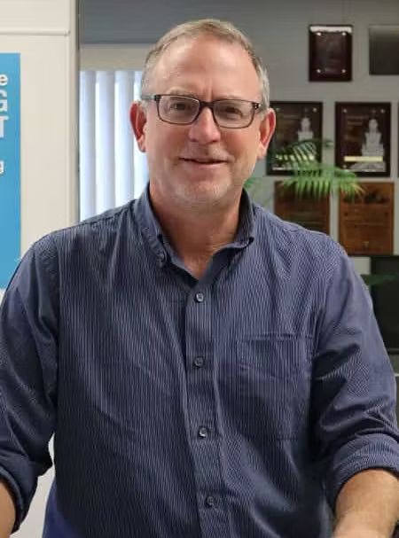 Man in a blue shirt and glasses standing in an office, smiling at the camera.