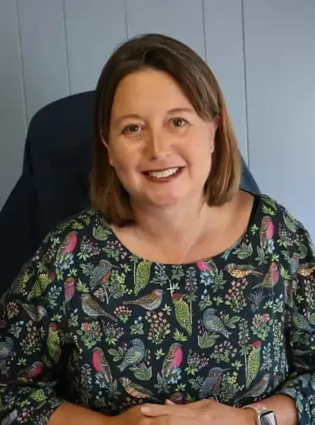 Smiling woman seated in a chair, wearing a dark floral top against a light wall.