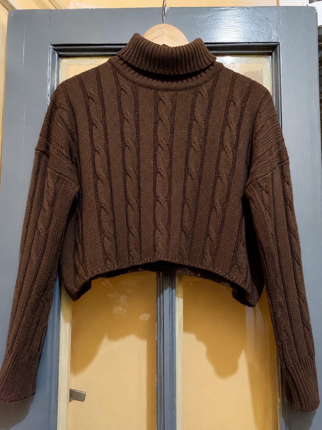 Mujer con jersey de cuello alto color crema y vaqueros de pie en una puerta con una mano en el bolsillo.