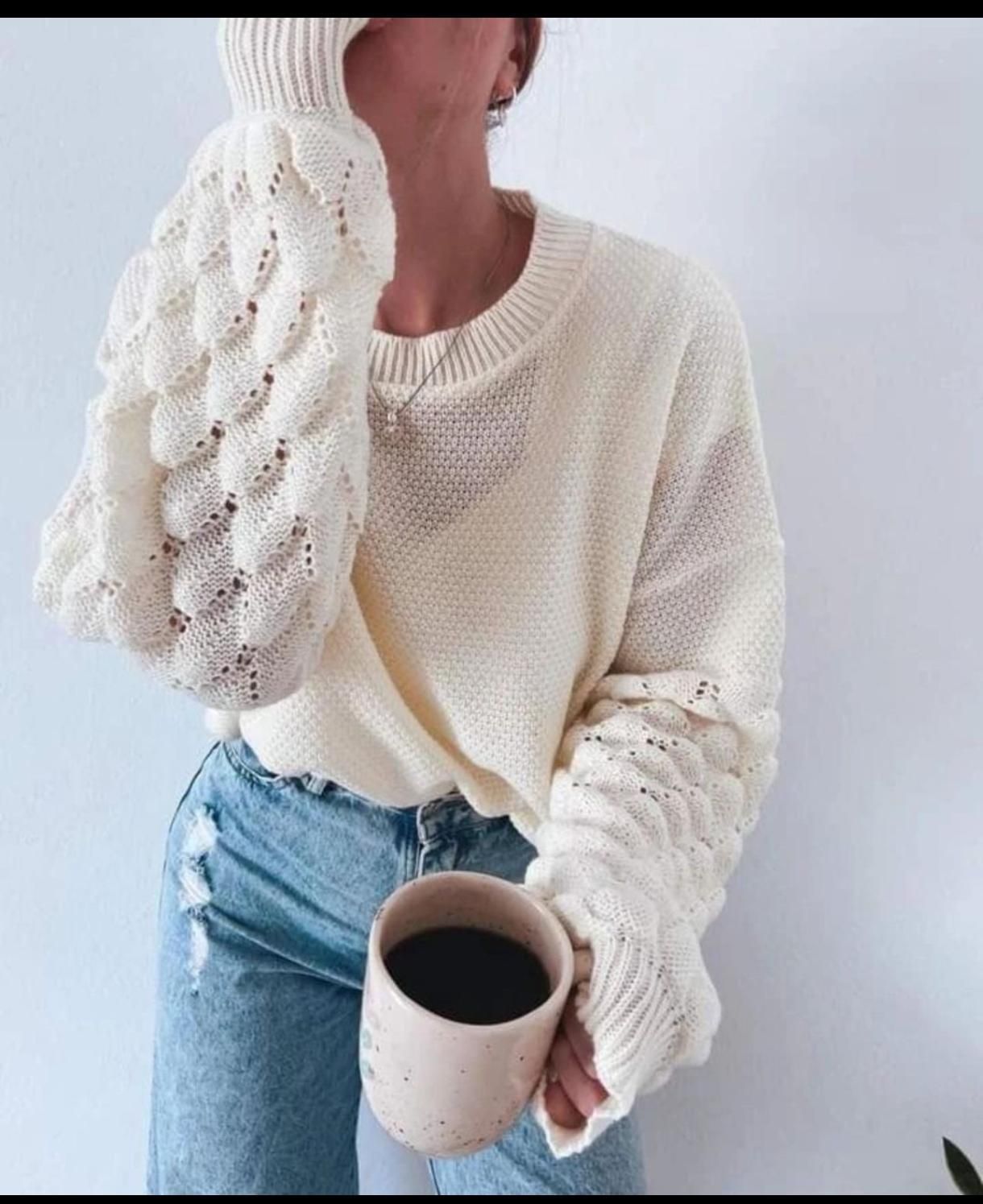 Persona con un suéter de punto color crema y jeans azules sosteniendo una taza de café sobre un fondo claro.