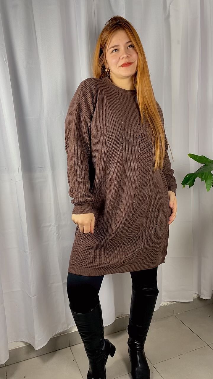 Mujer con un vestido de punto texturizado marrón y botas negras de pie en el interior de una casa junto a una cortina blanca.