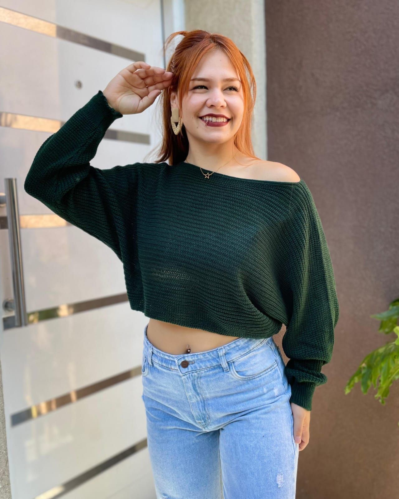 Mujer sonriente con suéter verde oscuro y jeans claros posando al aire libre junto a una pared.
