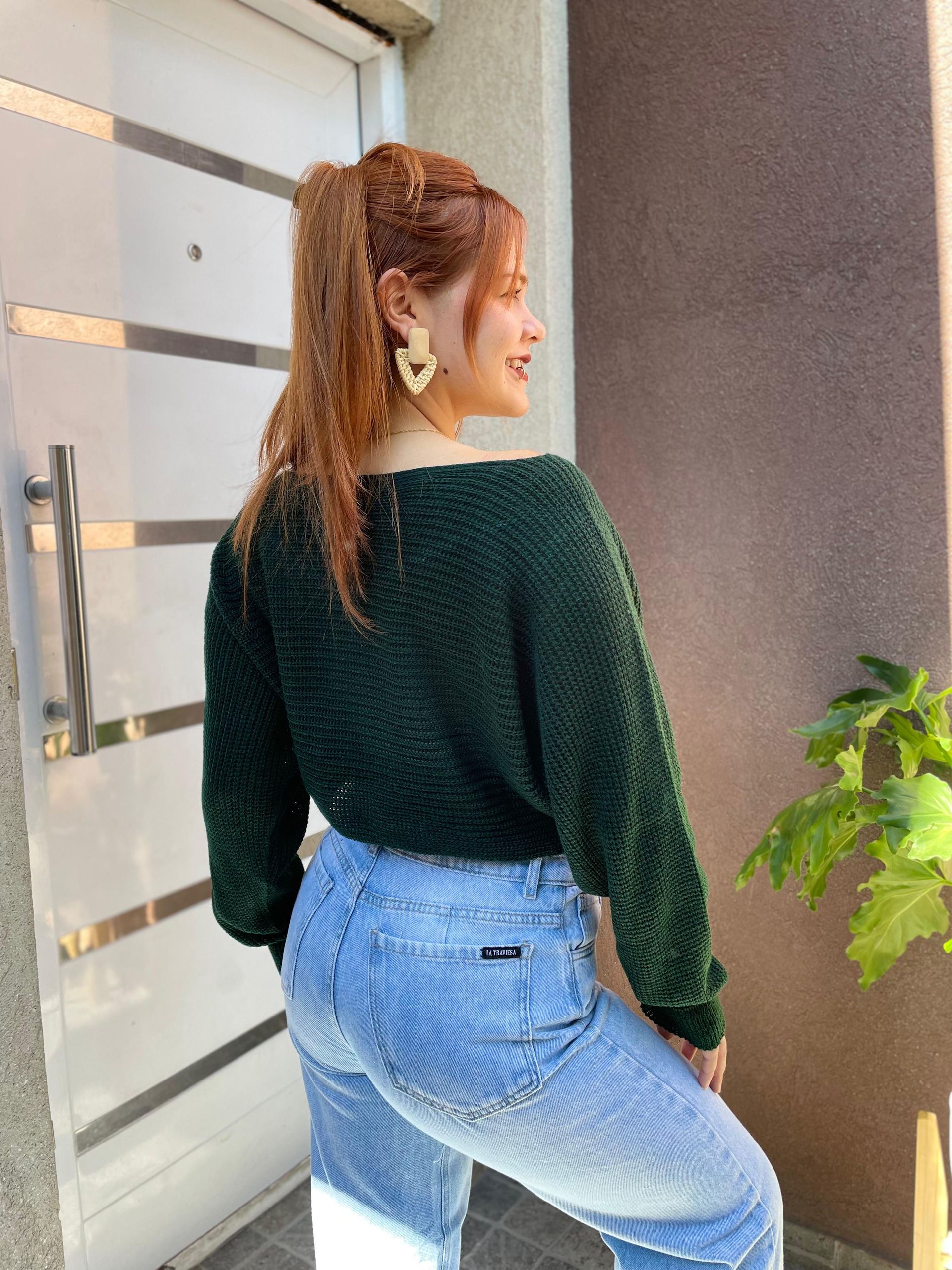 Mujer sonriente con suéter verde oscuro y jeans claros posando al aire libre junto a una pared.