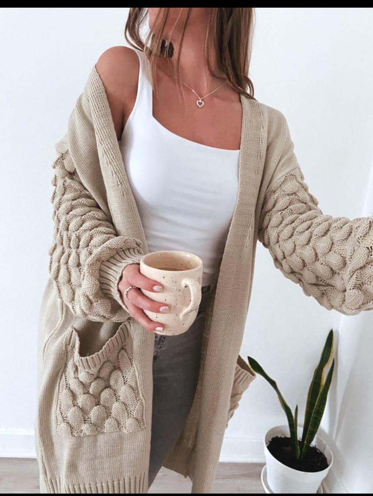 Una persona con un cárdigan beige sostiene una taza junto a una planta en maceta y lleva una blusa blanca.