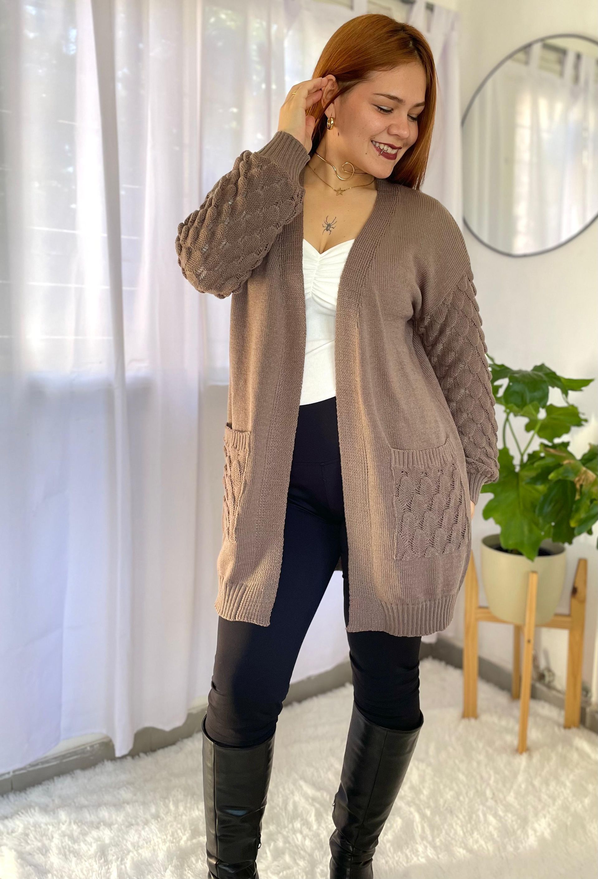 Una persona con un cárdigan beige sostiene una taza junto a una planta en maceta y lleva una blusa blanca.
