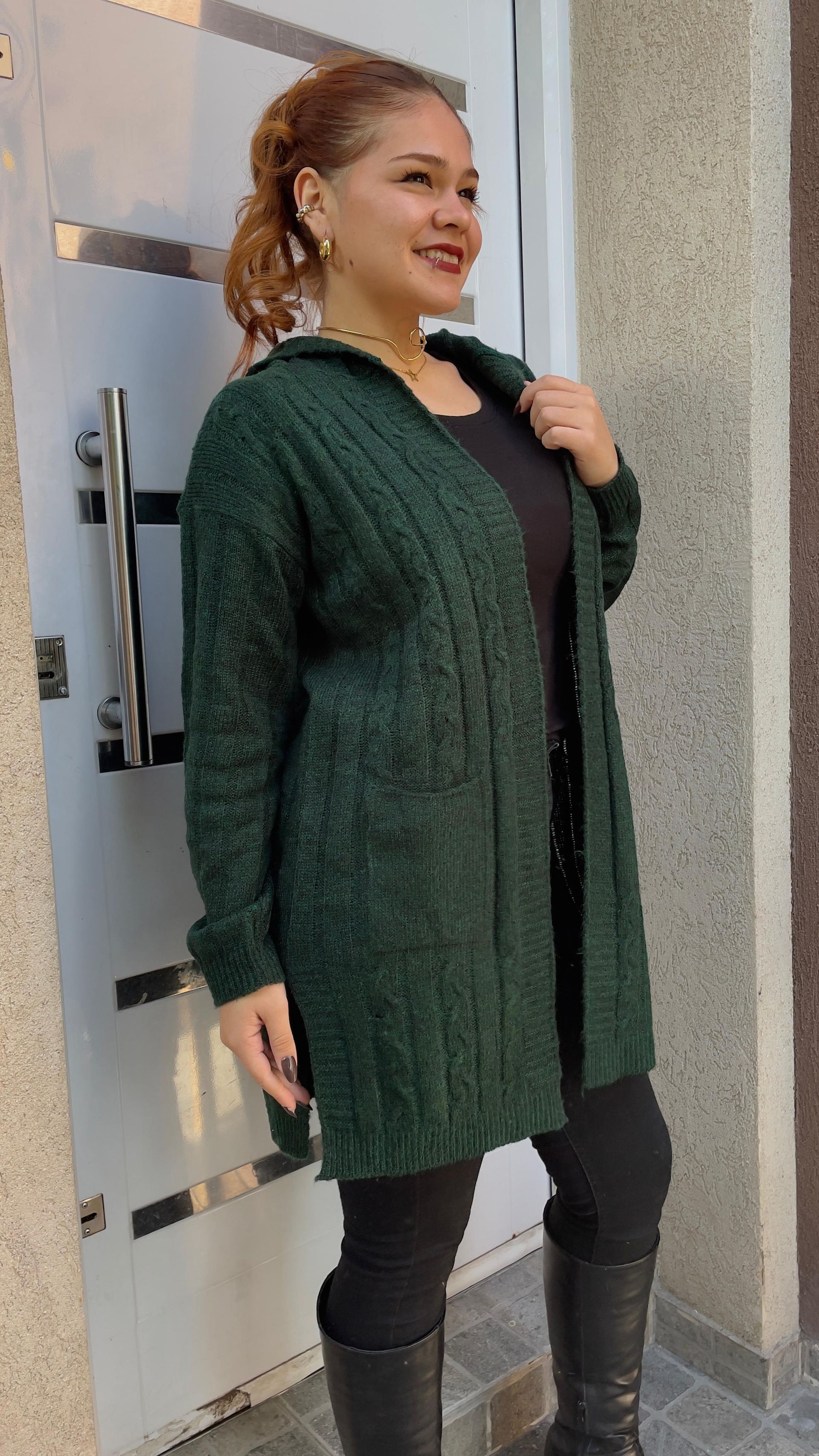 Persona con un abrigo verde oscuro de pie junto a una puerta blanca, sonriendo y levantando el cuello de la chaqueta.