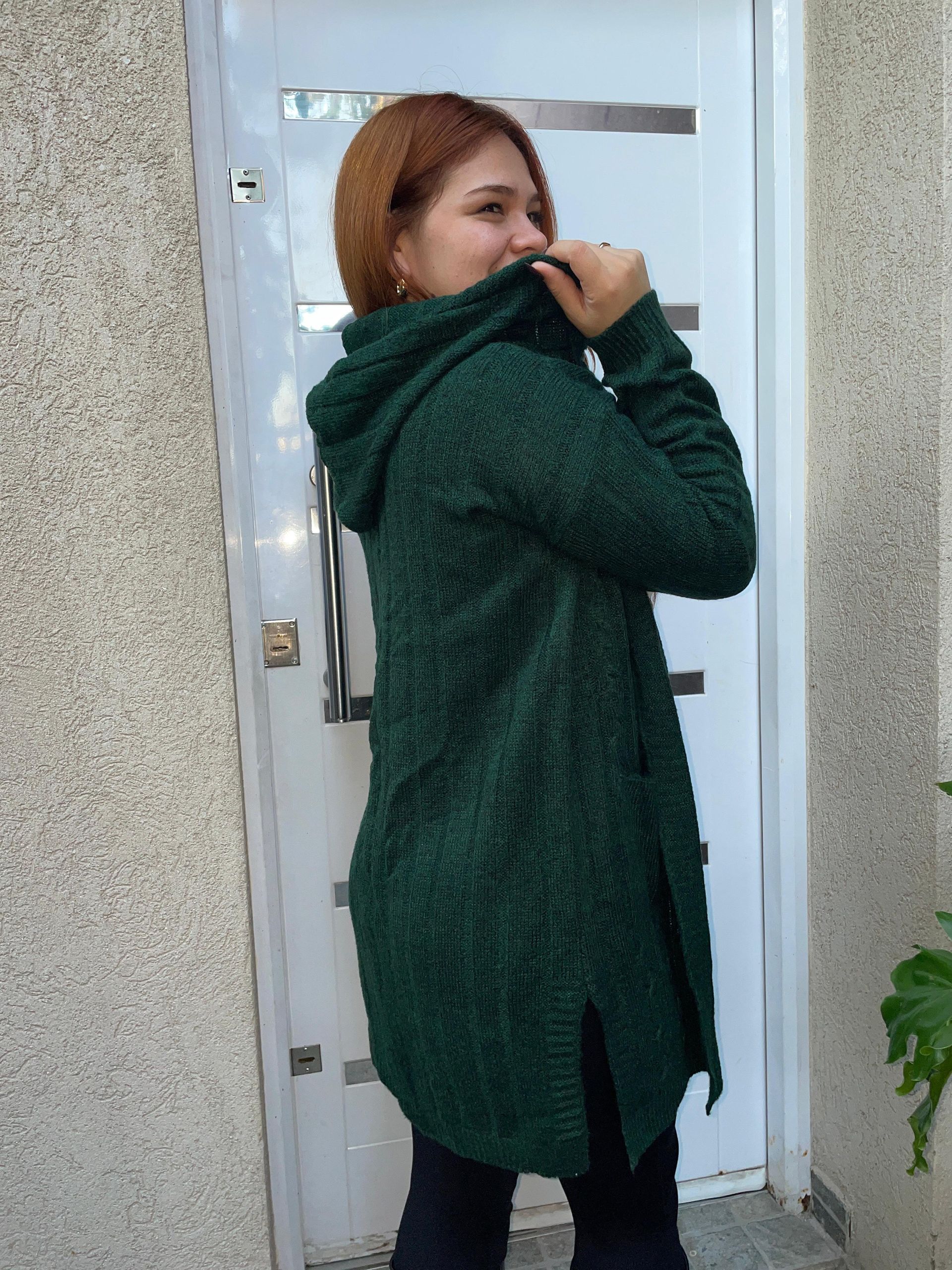 Persona con un abrigo verde oscuro de pie junto a una puerta blanca, sonriendo y levantando el cuello de la chaqueta.