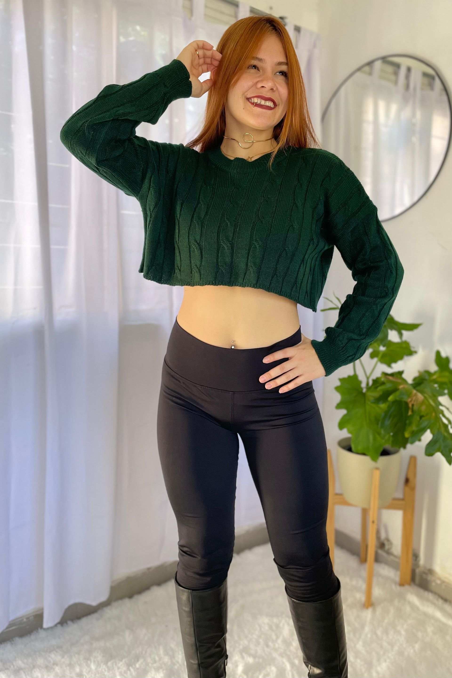 Mujer posando en el interior con un suéter corto verde y pantalones negros, de pie junto a una ventana y un espejo.