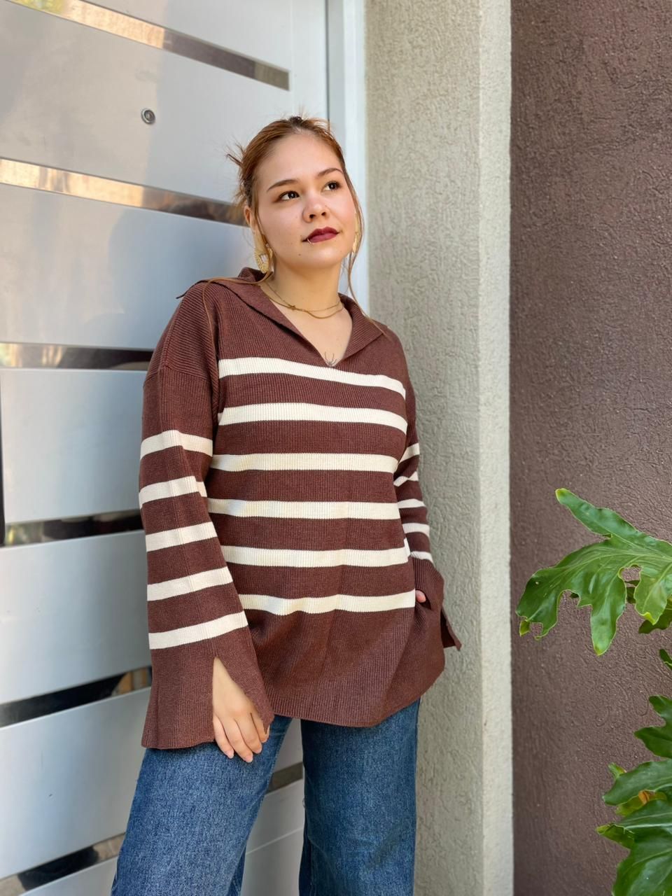 Mujer con suéter de rayas marrones y jeans de pie junto a una puerta al aire libre, mirando a la cámara.
